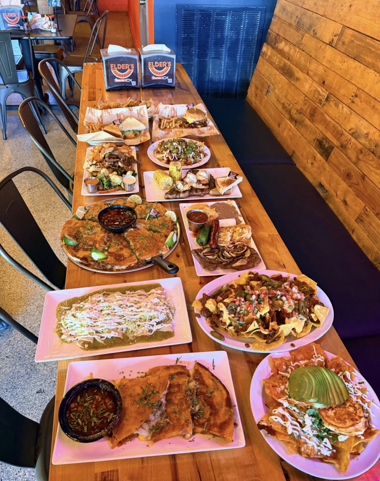 A long table spread of Elder's dishes: quesadillas, birria, nachos, steaks, sandwiches and tacos